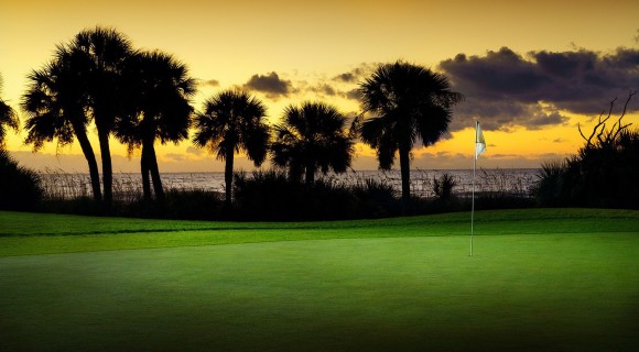 Golf course hole at sunrise with palm trees and ocean in background