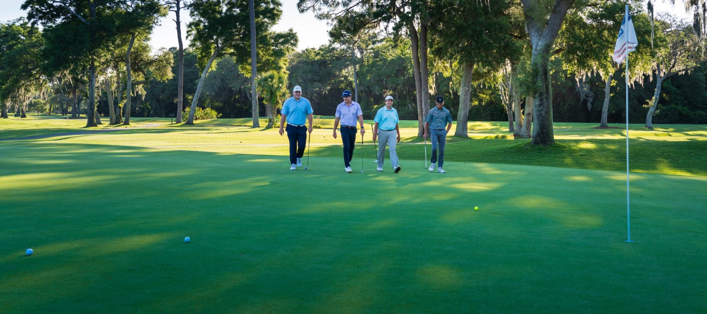 four male golfers walking towards golf balls and flag with sun and trees behind them