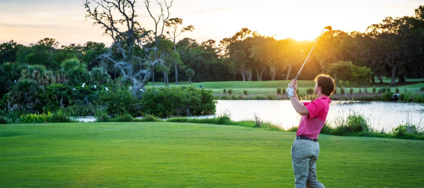 golfer in pink polo post swing with sun flare above golf club and water in background